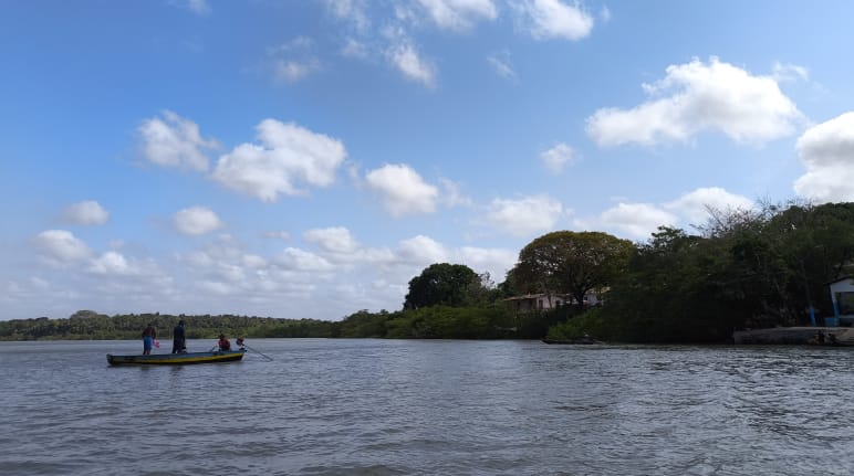 Pescadores artesanais da comunidade de Portinho, que faz parte da Resex Tauá-Mirim, mantêm viva a cultura e os saberes do território Sob o céu azul, um pequeno barco navega pelo mar com três pescadores. À direita estão pequenas casas no meio de uma floresta.
